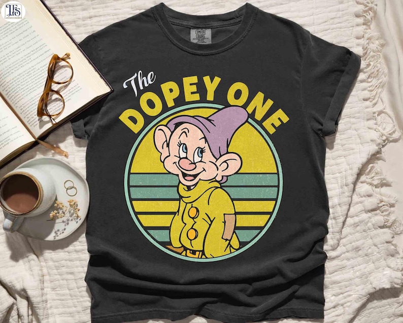 May include: Black t-shirt featuring a cartoon image of Dopey from Snow White. The graphic shows Dopey in a yellow outfit and purple hat, set against a yellow and blue striped circle. The text "The Dopey One" is above the character.