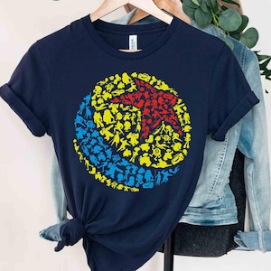 May include: Navy blue t-shirt featuring a colorful graphic of a ball composed of various character silhouettes. The design includes red, yellow, and blue figures, creating a playful and eye-catching design. The shirt is hanging on a wooden hanger.