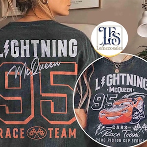 May include: Dark gray t-shirt featuring the text "LIGHTNING McQueen 95 RACE TEAM" in white and orange. The design includes a graphic of an orange race car. The shirt is part of the "2006 Piston Cup Series" collection.