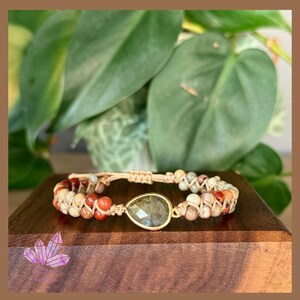 May include: A handcrafted bracelet with a central, teardrop-shaped gemstone in a gold-coloured setting. The bracelet is decorated with small, colourful beads in red, beige, and green, strung on a beige cord. The bracelet is displayed on a wooden block.