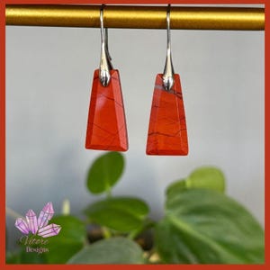 May include: A pair of red jasper earrings with silver hooks. The earrings are rectangular with a trapezoidal base. The red stone features dark veins. The earrings are suspended from a gold bar. Each earring measures approximately 3 cm in length.