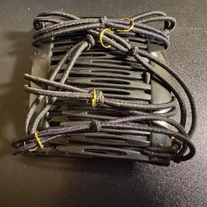May include: Black hair combs with elastic bands and gold-colored accents. The combs are stacked and wrapped with black elastic bands, secured with small gold-colored rings. These hair accessories are designed to hold hair in place.