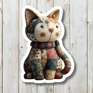 May include: A fabric cat sticker with a patchwork design. The cat has a white face, green eyes, and a pink nose. The body is made of various patterned fabrics in shades of blue, red, and beige. It is wearing a patterned scarf.