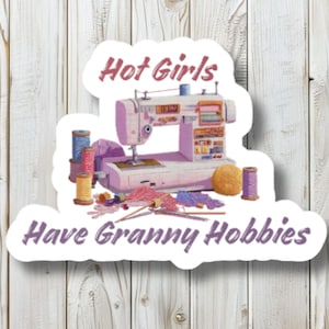 Könnte beinhalten: Ein Aufkleber mit einer Nähmaschine, Garnrollen, Wolle und Stricknadeln. Der Text lautet "Hot Girls Have Granny Hobbies" in lila Schrift. Der Aufkleber hat einen weißen Rand und befindet sich auf einem Holzmaserungshintergrund.