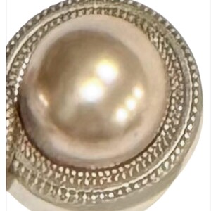 May include: A close-up of a silver-toned button with a large, lustrous, pearlescent center. The button has a detailed, textured border. The overall design is elegant and classic, suitable for various garments.