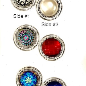 Puede incluir: Cinco botones decorativos redondos, de tono plateado, con diferentes diseños. Los botones tienen un borde elevado con cuentas. Los diseños incluyen un patrón geométrico en blanco y negro, una perla blanca, una mandala multicolor, un patrón de cuadros rojos y una mandala azul y verde.