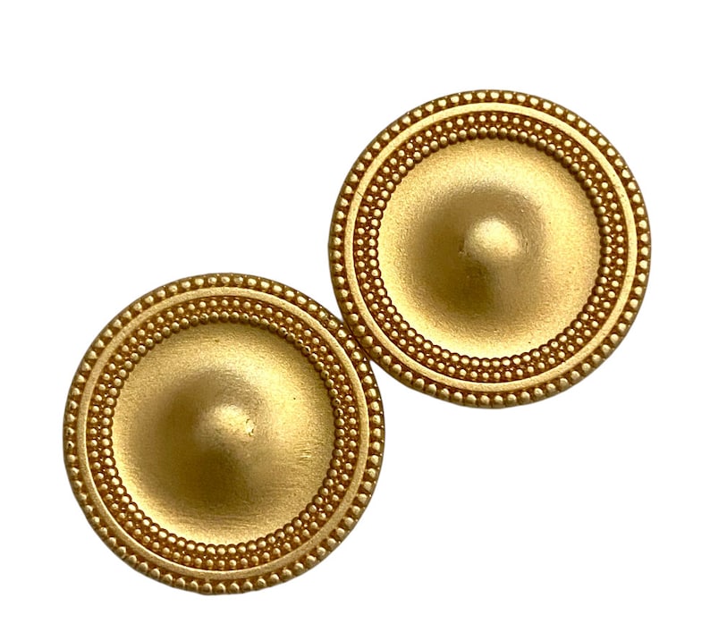 May include: A pair of gold-toned metal earrings with a circular design. The earrings have a raised, domed centre and a beaded border.