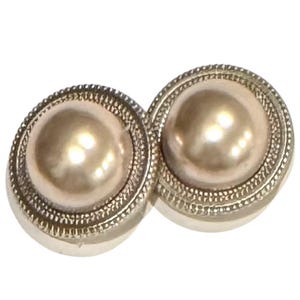May include: A pair of round, pearl-like earrings. Each earring features a large, light-colored, domed center surrounded by a silver-colored, textured border. The earrings are set against a white background.