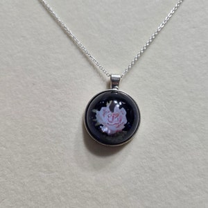 May include: A silver necklace with a round pendant featuring a pink rose on a dark blue background. The pendant is encased in a silver-colored metal frame. The chain is delicate and hangs from the pendant.
