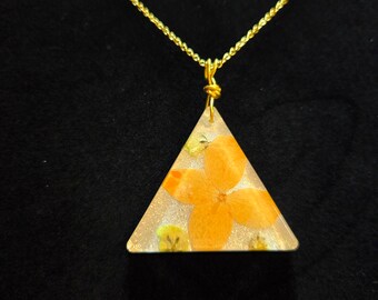 May include: A gold-colored chain necklace with a triangular pendant. The pendant is clear with an orange flower and small yellow flowers encased inside. The background is black.