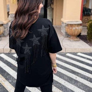 May include: A black oversized t-shirt with a back design of four star-shaped embellishments with dangling fringe. The shirt is worn with black leggings and white boots.