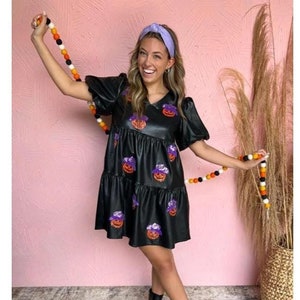 Puede incluir: Un vestido negro de cuero sintético corto con estampado de calabaza y falda con volantes. El vestido tiene mangas cortas abullonadas y un escote en V. La modelo lleva una diadema de color morado claro y botines negros.