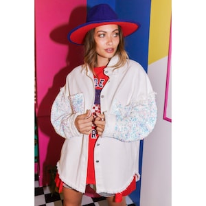 May include: White denim jacket with sequined sleeves. The jacket has a button-up front and a relaxed fit. The model is wearing a red and blue striped shirt underneath the jacket.