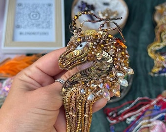Puede incluir: Un broche con forma de dragón elaborado con lentejuelas, cuentas y cristales en tonos oro, bronce y marrón. La pieza presenta una cabeza detallada con un ojo blanco, un cuerpo texturizado y largos flecos de cadenas doradas colgantes.