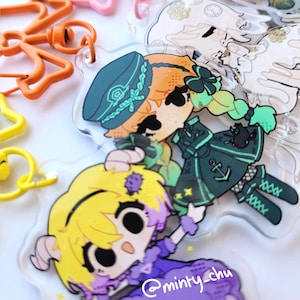 May include: Clear acrylic charms depicting anime-style characters in various outfits. The charms have a glossy finish and are in shades of yellow, orange, green, and purple. The text "@minty_chu" is visible on one of the charms. The charms are attached to colourful keychains.