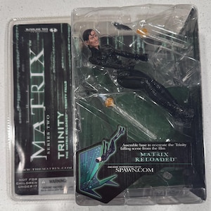 May include: A Trinity action figure from the Matrix Reloaded film, packaged in a clear plastic case. The figure is in a dynamic pose, with the words "MATRIX RELOADED" and "TRINITY" visible on the packaging. The packaging also includes the text "McFarlane Toys".