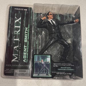 Puede incluir: Figura de acción de Agent Smith de la serie Matrix Revolutions, empaquetada en una funda de plástico transparente. La figura está en una pose dinámica, con un traje negro y gafas de sol. El embalaje incluye el logotipo de Matrix y el texto: "Agent Smith".