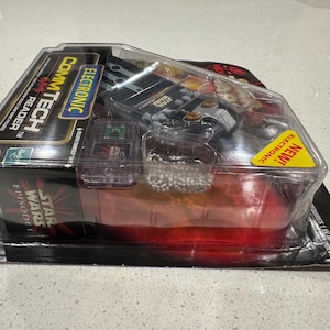 Puede incluir: Un juguete Star Wars Episodio I CommTech Reader en su embalaje original. El embalaje es negro con detalles en rojo y amarillo, y el juguete es visible a través de una ventana de plástico transparente. Incluye el texto "ELECTRONIC" y "NEW!"
