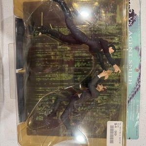 Puede incluir: Un juego de figuras de acción empaquetado con dos figuras en poses dinámicas, vestidas con trajes negros. Las figuras están en un paquete de plástico transparente con un respaldo dorado. El texto "NEO VS. AGENT SMITH" es visible en el embalaje. El set parece ser de una película.