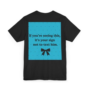 Pode incluir: Camiseta preta com um gráfico quadrado turquesa com bolinhas brancas e o texto "If you're seeing this, it's your sign not to text him." Um laço preto está abaixo.