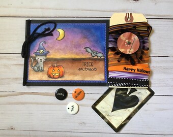 May include: A Halloween themed craft kit with a black and white illustration of a cat wearing a witch's hat and a pumpkin. The kit includes a card with the words "Trick or Treat", a tag with the words "Happy Halloween", and a variety of ribbons and trims in orange, black, and purple.