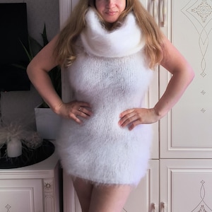 May include: A white sleeveless mini dress crafted from a fuzzy, mohair-style knit fabric. The garment features a prominent, thick, oversized cowl neckline that drapes around the neck and a fitted silhouette that ends at the upper thigh.