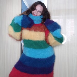 May include: A long, oversized rainbow-striped sweater with a high turtleneck. The sweater features horizontal bands of red, orange, yellow, green, blue, and purple. The sleeves are long and voluminous, and the material appears soft and fuzzy.