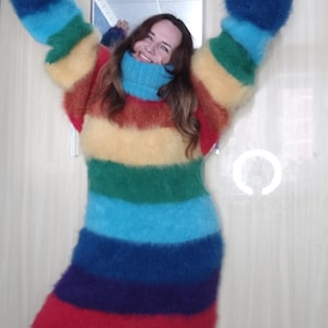 May include: A long, rainbow-striped sweater dress with a high turtleneck. The dress features horizontal bands of red, orange, yellow, green, blue, and purple. The sleeves are long and oversized, and the dress appears to be made of a soft, fuzzy material.