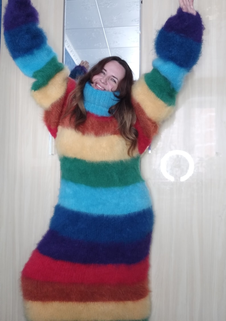 May include: A long, rainbow-striped sweater dress with a high turtleneck. The dress features horizontal bands of red, orange, yellow, green, blue, and purple. The sleeves are long and oversized, and the dress appears to be made of a soft, fuzzy material.