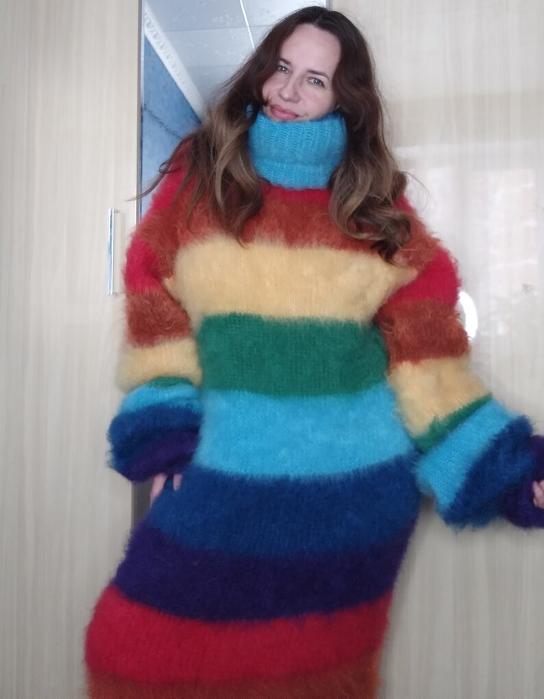 May include: A long, rainbow-striped sweater dress with a high, turquoise turtleneck. The dress features horizontal bands of red, orange, yellow, green, blue, and purple. The sleeves are long and fluffy, matching the soft, textured appearance of the dress.
