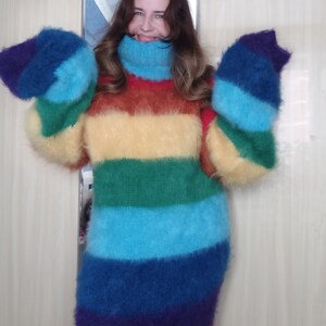 May include: A long, rainbow-striped sweater dress with a high turtleneck and long sleeves. The dress features horizontal bands of red, purple, blue, green, yellow, and orange. The fluffy material creates a cozy and vibrant look.