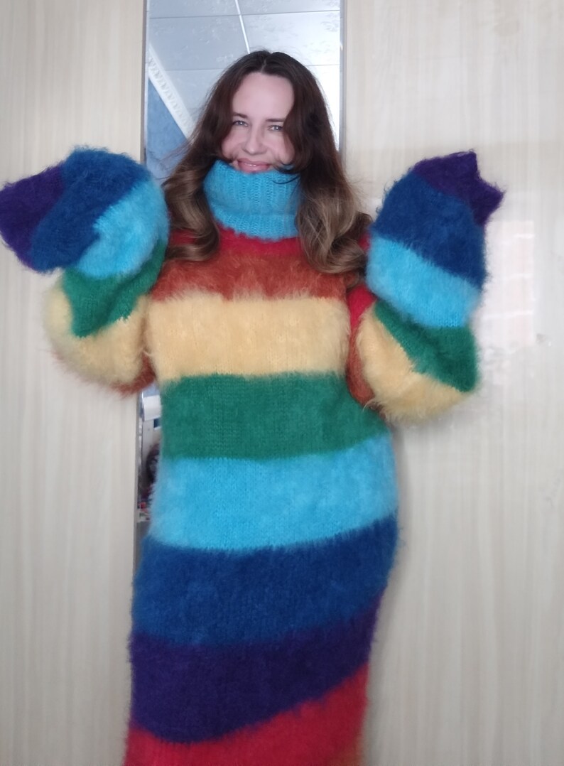 May include: A long, rainbow-striped sweater dress with a high turtleneck and long sleeves. The dress features horizontal bands of red, purple, blue, green, yellow, and orange. The fluffy material creates a cozy and vibrant look.