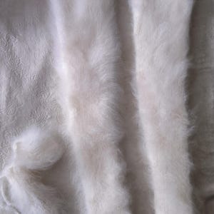 Peut inclure: Une paire de jambières longues, blanches et duveteuses avec une texture douce et brossée. À côté des jambières se trouve une petite pelote de laine blanche assortie. Les articles sont fabriqués dans une matière légère et aérée.