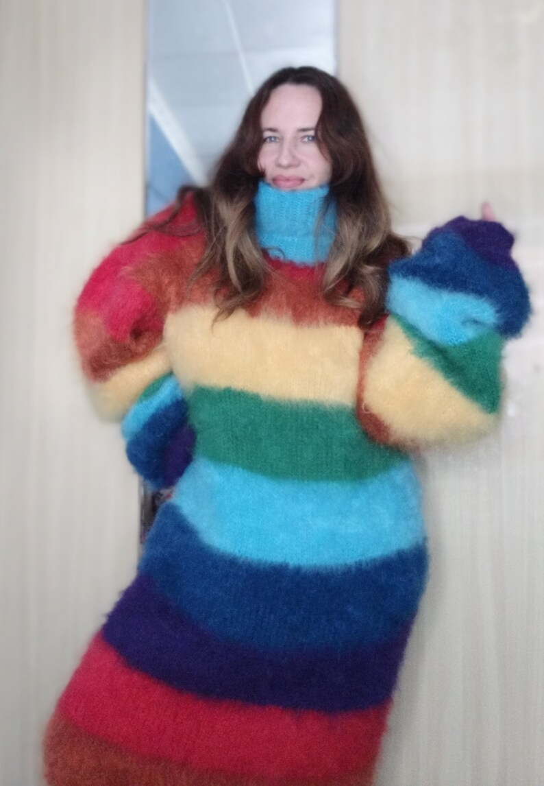 May include: A long-sleeved, rainbow-striped sweater with a turtleneck. The sweater features bands of red, orange, yellow, green, blue, and purple. The sleeves are long and puffy, and the sweater appears to be made of a soft, fuzzy material.