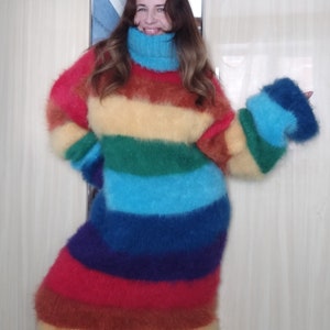 May include: A long, rainbow-striped sweater with a high, light blue turtleneck. The sweater features horizontal stripes in red, orange, yellow, green, blue, and purple. The sleeves are long and flared, matching the sweater's fluffy texture.