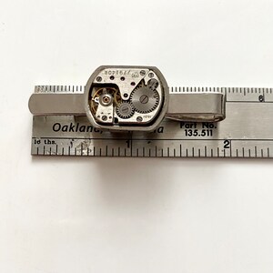 Puede incluir: Pinza de corbata plateada con un mecanismo de relojer&iacute;a visible. La pinza est&aacute; sobre una regla con el texto "Part No. 135.511" y "Oakland, Ca".
