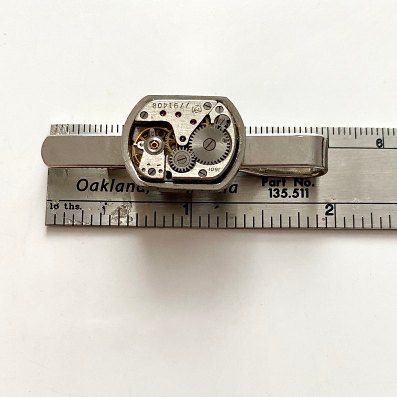 Puede incluir: Pinza de corbata plateada con un mecanismo de relojer&iacute;a visible. La pinza est&aacute; sobre una regla con el texto "Part No. 135.511" y "Oakland, Ca".