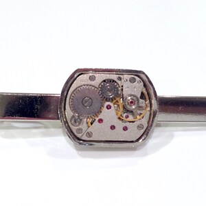 Puede incluir: Pinza de corbata plateada con una caja octogonal transparente que revela un movimiento de reloj plateado y dorado con joyas rojas.