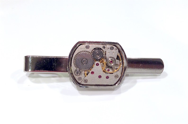 Puede incluir: Pinza de corbata plateada con una caja octogonal transparente que revela un movimiento de reloj plateado y dorado con joyas rojas.