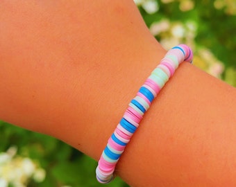May include: A colorful beaded bracelet with pink, blue, and white beads. The bracelet is on a person's wrist.