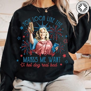 May include: Black sweatshirt with a graphic of a woman dressed as the Statue of Liberty holding a hot dog and a book. Text reads "You look like the 4th of July makes me want hot dog real bad."