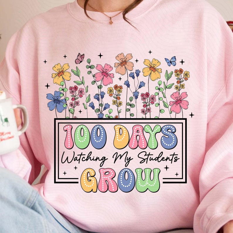 Pode incluir: Camisola rosa claro com um design floral e o texto "100 Days Watching My Students Grow" num ret&acirc;ngulo preto. O design floral inclui flores coloridas e borboletas. A camisola &eacute; de cor rosa claro.
