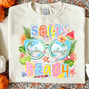 May include: Cream sweatshirt featuring the words "Salty Beach" in a colourful design. The graphic includes blue sunglasses reflecting a beach scene, tropical flowers, fruit slices, and seashells. A fun, summery design.