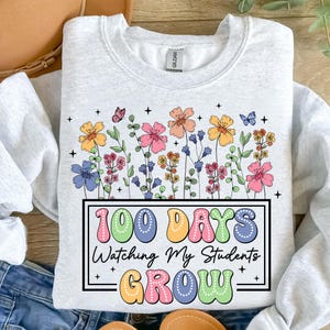 Pode incluir: Um moletom cinza claro com um design floral colorido e borboletas. O texto "100 DAYS Watching My Students GROW" &eacute; exibido em uma moldura retangular, usando cores pastel. A etiqueta da marca Gildan &eacute; vis&iacute;vel.