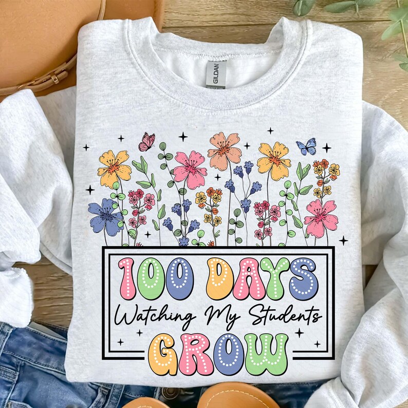 Pode incluir: Um moletom cinza claro com um design floral colorido e borboletas. O texto "100 DAYS Watching My Students GROW" &eacute; exibido em uma moldura retangular, usando cores pastel. A etiqueta da marca Gildan &eacute; vis&iacute;vel.