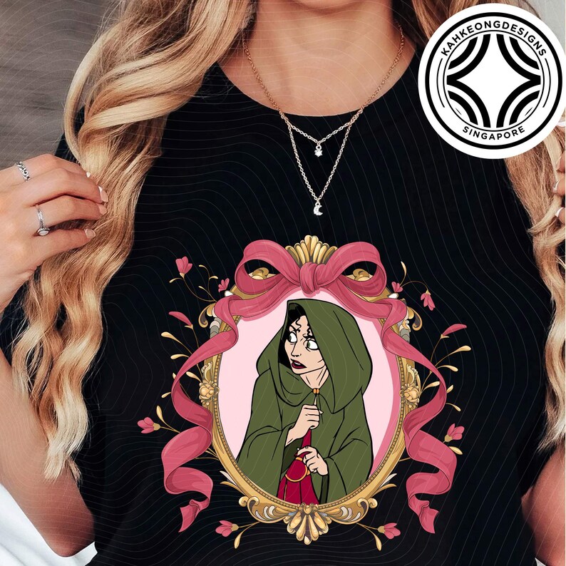 Puede incluir: Camiseta negra con una ilustraci&oacute;n enmarcada de un personaje con una capa con capucha verde, sosteniendo una bolsa roja. El marco es dorado con una cinta rosa y detalles florales. Se ve un collar dorado con un dije de estrella y luna.