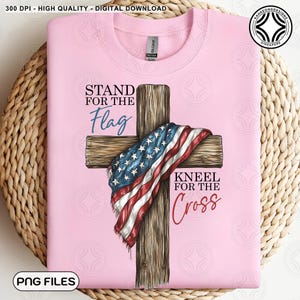 May include: A pale pink sweatshirt with the text "STAND FOR THE Flag, KNEEL FOR THE Cross". The design features a wooden cross draped with an American flag. The words are in a mix of fonts.