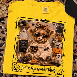 May include: A bright yellow t-shirt featuring a graphic of a fluffy Highland cow wearing heart-shaped sunglasses and a patterned headband. The cow holds a jack-o'-lantern mug, surrounded by pumpkins, ghosts, and books with the text "just a few spooky things".