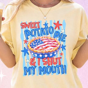 May include: Yellow t-shirt with a graphic of a sweet potato pie decorated with the American flag. The text on the shirt reads "Sweet Potato Pie & I Shut My Mouth" in red and blue lettering, with star accents.
