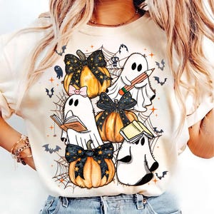 May include: A cream-colored t-shirt featuring a graphic print of three orange pumpkins with black star-patterned bows, three white ghosts, spiderwebs, bats, and small orange stars. One ghost holds a pencil, one reads a book, and one wears a pink bow.